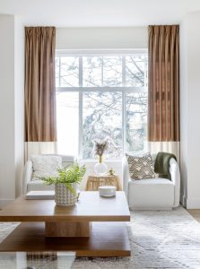 A South Surrey living room in The Loop townhome 'Highline' floor plan, featuring two white armchairs, a large window with two-tone curtains, and a two-tiered wood coffee table.